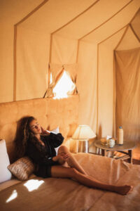 Sensual glamour portrait of Asian woman in interior of modern luxury glamping tent camp in Morocco. Relaxing on bed. Eco design concept, natural and wooden elements in style.