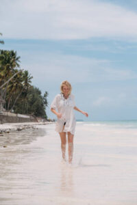 Happy woman having fun, enjoying summer, running joyfully on tropical beach.She is wearing white beach tunic on vacations on perfect Paje beach, Zanzibar, Tanzania.