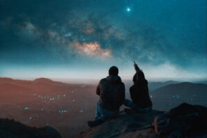 silhouette of a couple sitting on top of a hill looking at the stars over the city