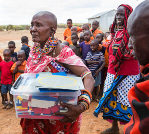 Amboseli, Kenya - November 2, Senior maasai woman with box of Lego wached by children. They are standing outside a primary school and some of them have school uniforms. The adult women are dressed in traditional kangas.