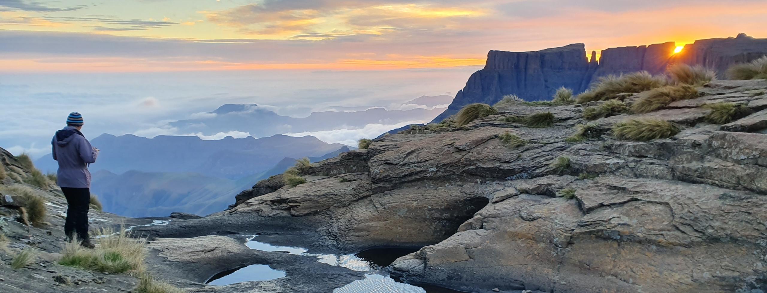 A person stands atop the Drakensberg Mountains, gazing at the sun as it sets over the breathtaking landscape.