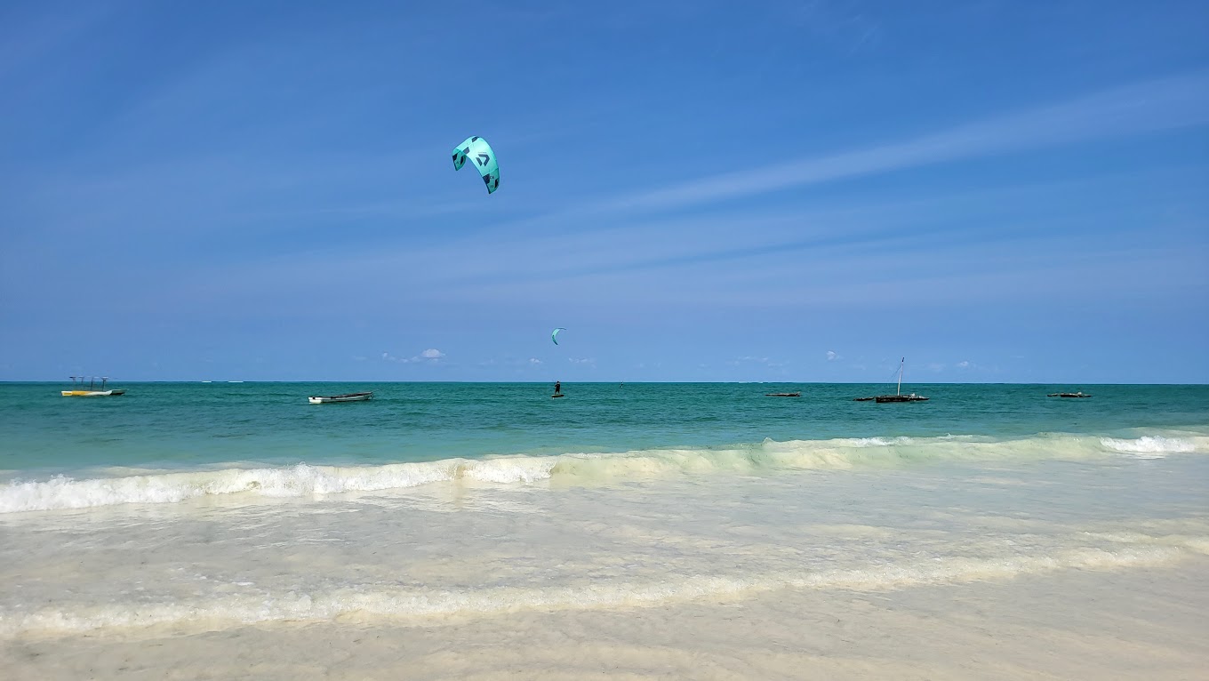 A person at Paje Beach joyfully flies a colorful kite against a clear blue sky, with waves gently lapping the shore.
