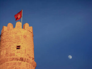 Tower of the Ribat fortress in Monastir and Tunisian Flag with the moon and blue clear sky on the background. Sunset hour