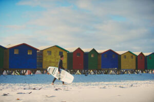 Surfer walking with surfboard with colourful beach huts at Muizenberg Beach Cape Town South Africa