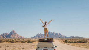 Young woman with long hair feeling freedom staying on the roof tent of the 4X4 camper truck, contemplating the road trip along the dramatic mountain landscape 