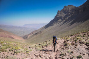 Female Hiker walking up a mountain pass with backpack on her back on a single footpath up a mountain with a valley and a clear blue sky behind Atlas Mountain Toubkal Morocco